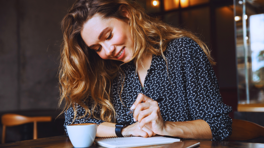 Frau sitzt in einem Café und schreibt entspannt in ein Notizbuch