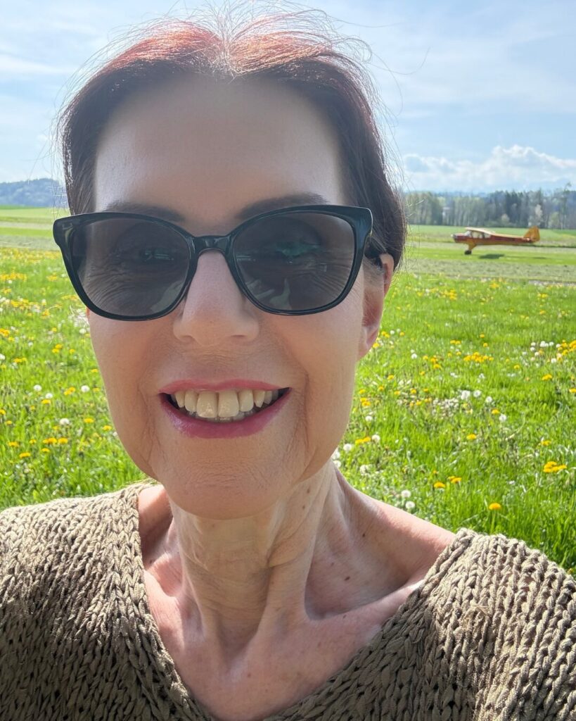 Gabriele Danners mit Sonnenbrille auf einer Wiese mit Blumen und Flugzeug im Hintergrund
